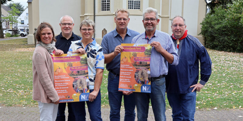 Zwei Frauen und vier Männer halten Veranstaltungsplakate hoch, im Hintergrund die Dorfkirche. Foto: Stadt Neukirchen-Vluyn/Sabrina Daubenspeck