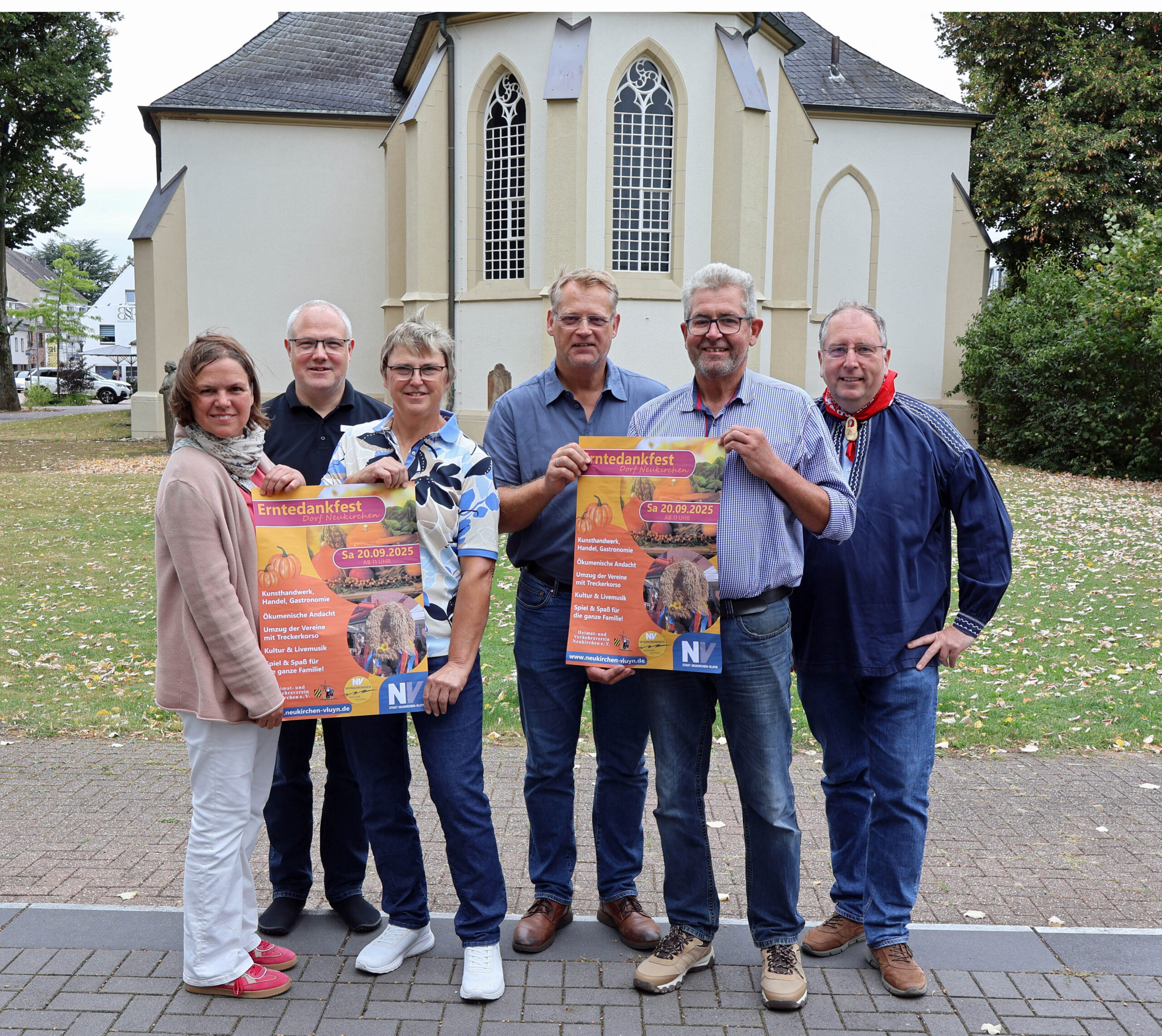 Zwei Frauen und vier Männer halten Veranstaltungsplakate hoch, im Hintergrund die Dorfkirche. Foto: Stadt Neukirchen-Vluyn/Sabrina Daubenspeck