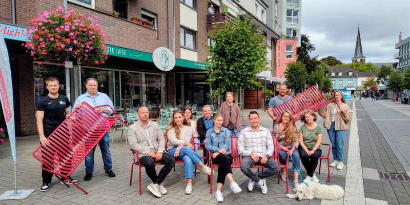 Eine Gruppe aus 13 Menschen und einem Hund sind auf dem Vluyner Platz. Einige sitzen auf roten Bänken, andere halten rote Bänke hoch.