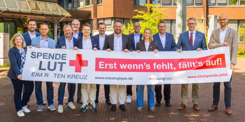 Frauen und Männer stehen hinter einem Banner und werben für den Blutspendertag 2026